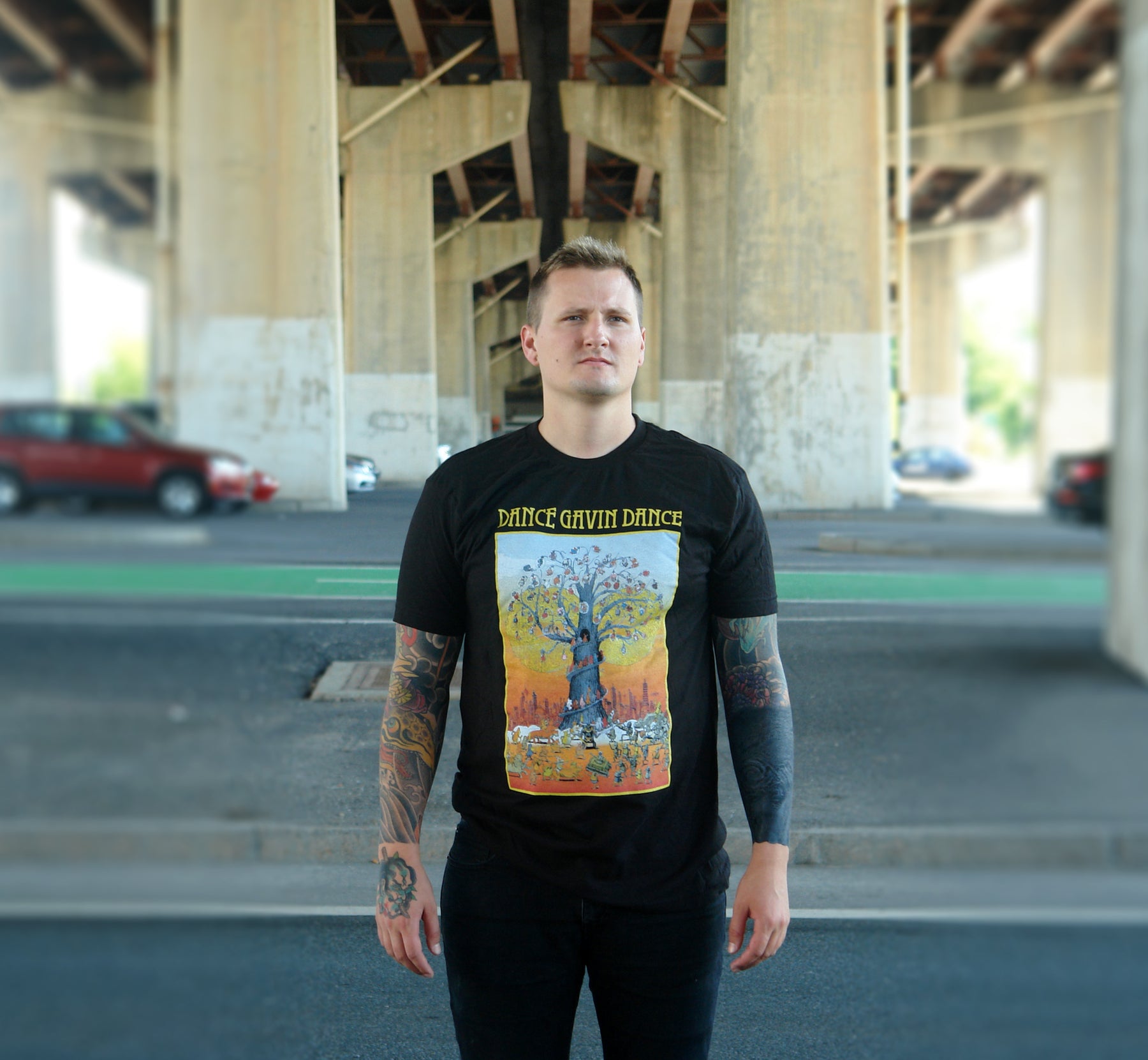 white man with short hair and tattoos on both arms standing under bridge wearing black pants and black tee shirt. black tee shirt has full chest print with yellow print on top that says dance gavin dance and a rectangle below that shows a multicolored tree with several characters surrounding it.