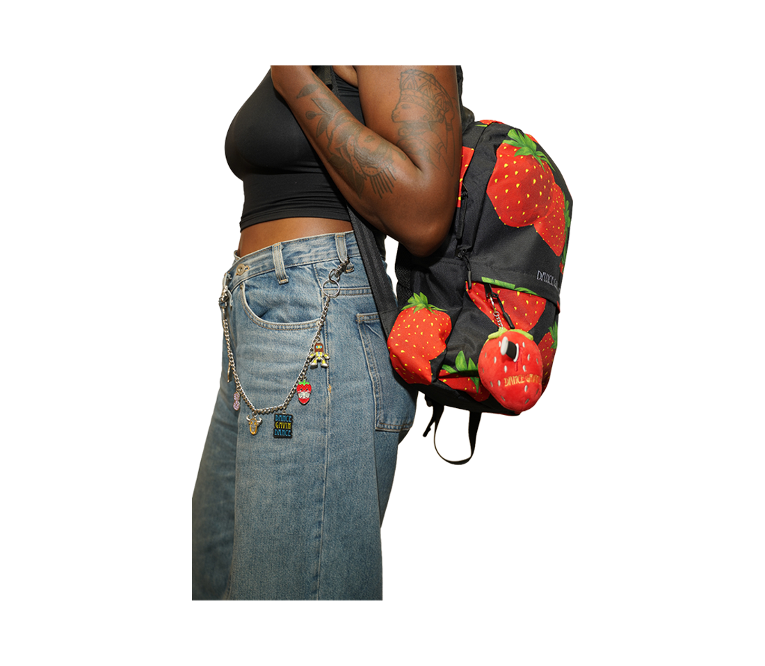 A person wearing a black crop top and light-wash denim jeans with a chain belt and a strawberry-patterned backpack, shown from the waist up against a plain white background.