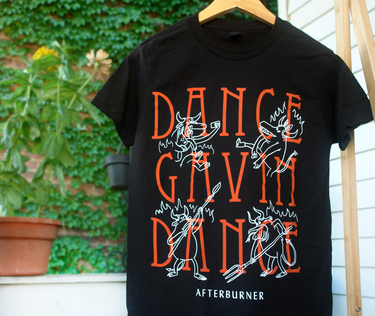 hanging black tee shirt in front of green leaves with full chest print that says dance gavin dance in stacked orange print and has four white outlined characters dispersed around and says afterburner on the bottom