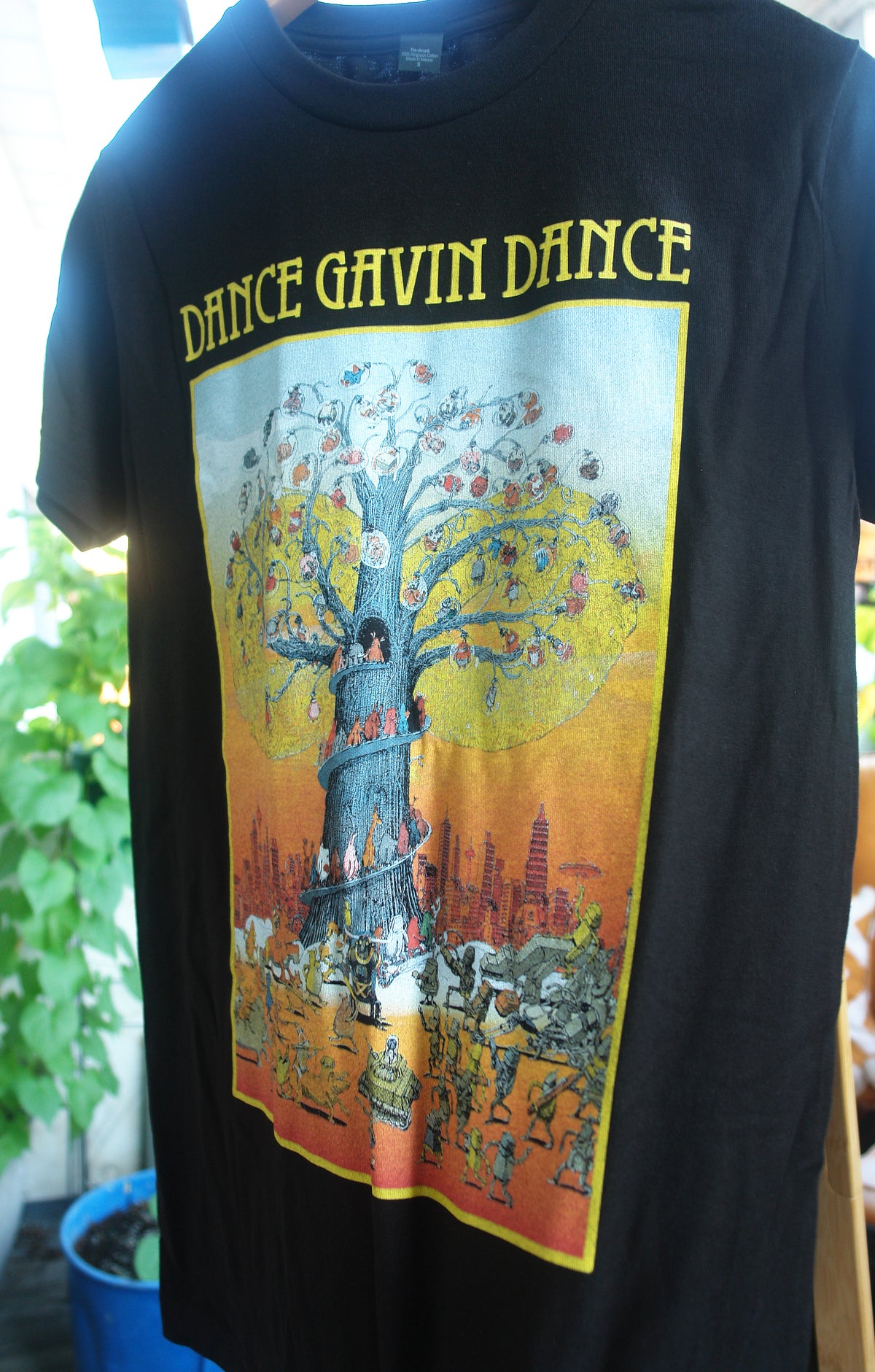 close up of black tee shirt in front of green leaves black tee shirt has full chest print with yellow print on top that says dance gavin dance and a rectangle below that shows a multicolored tree with several characters surrounding it.