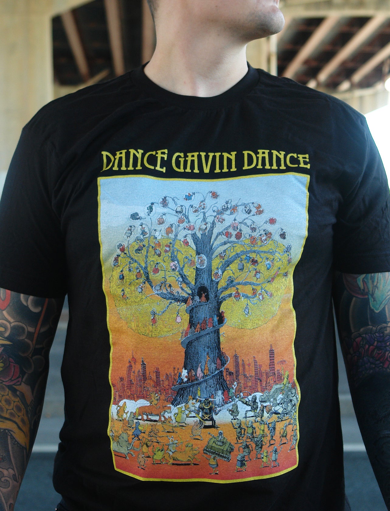 close up of white man with tattoos on both arms standing under bridge wearing black tee shirt. black tee shirt has full chest print with yellow print on top that says dance gavin dance and a rectangle below that shows a multicolored tree with several characters surrounding it.