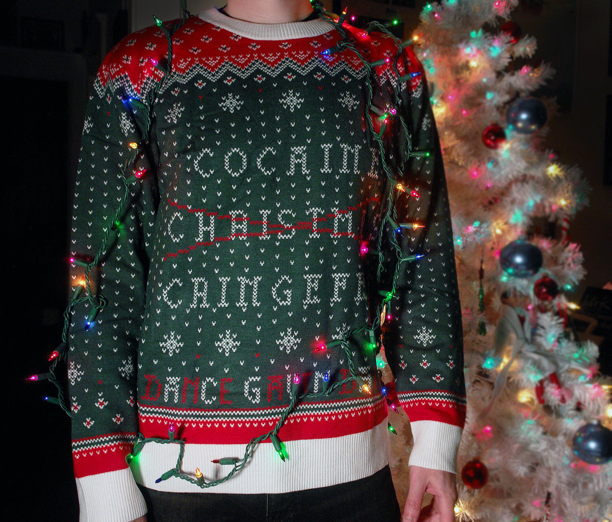 image of a custom holiday sweater on a man in front of a white christmas tree with lights drapped over him. the sweater is red, green & white and has the words cocaine, christmas, cringefest in the center and dance gavin dance across the bottom