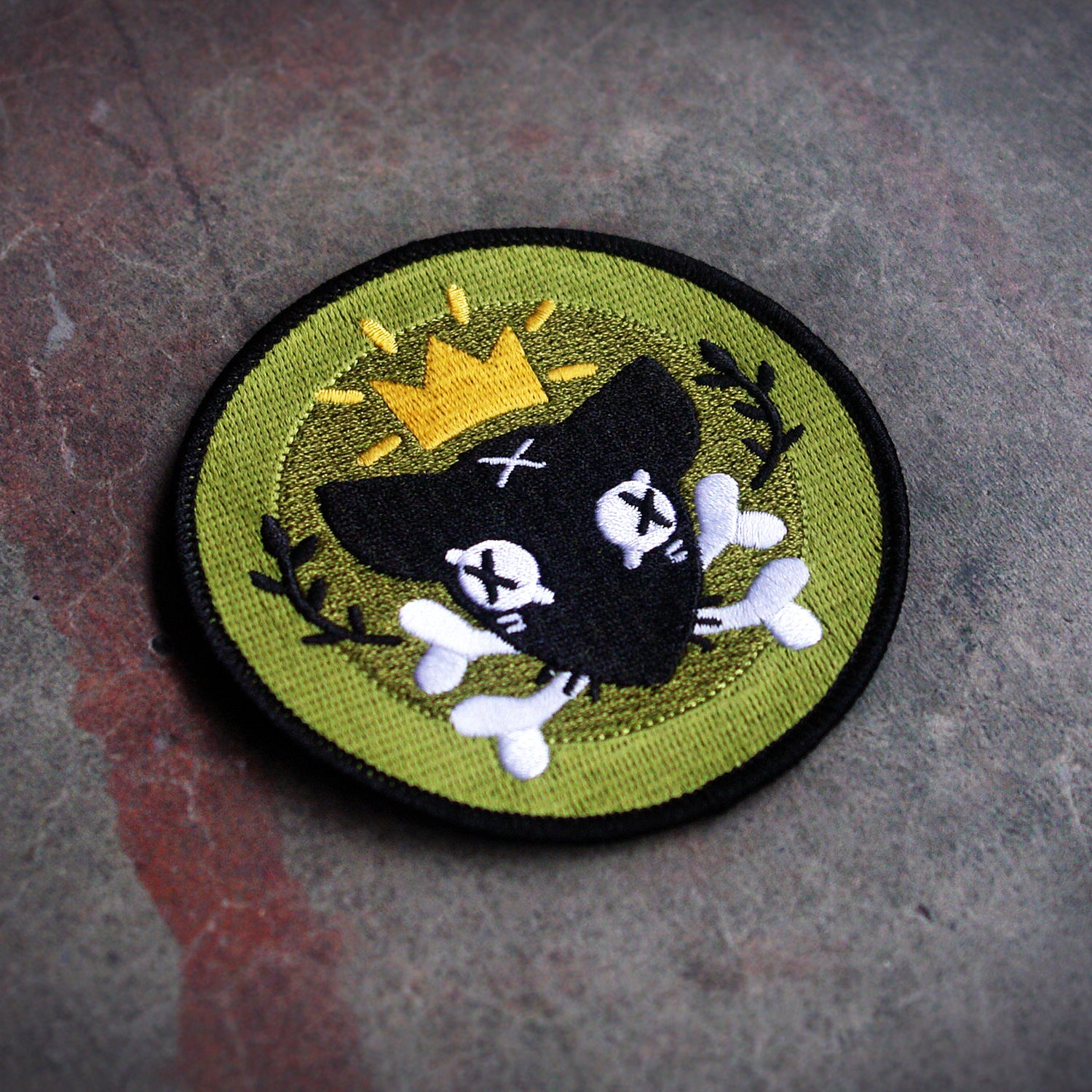 image of a circle patch mock up on a concrete background. black rim of circle with dead black rat with black x's as eyes and white bones in mouth wearing a gold crown framed with black laurels and a two tone green backgound.